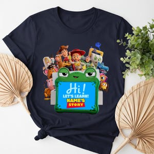 May include: Navy blue t-shirt with a vibrant graphic of animated characters. The design includes a frog-shaped frame with the word "Hi!" and the text "LET'S LEARN! NAME'S STORY".