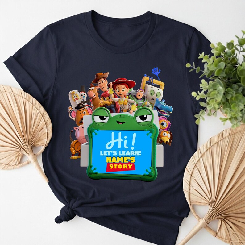 May include: Navy blue t-shirt with a vibrant graphic of animated characters. The design includes a frog-shaped frame with the word "Hi!" and the text "LET'S LEARN! NAME'S STORY".