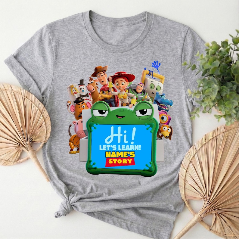 May include: A grey t-shirt with a vibrant graphic of animated characters. The design includes a green frog-shaped frame with the word "Hi!" and the text "LET'S LEARN! NAME'S STORY".