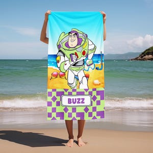 May include: A rectangular beach towel featuring a graphic of the character Buzz Lightyear on a sandy beach with a blue sky. The towel includes a snorkel, sunglasses, and a seashell. The bottom section has a purple and green checkered pattern with a white rectangular label containing the text BUZZ in purple capital letters.