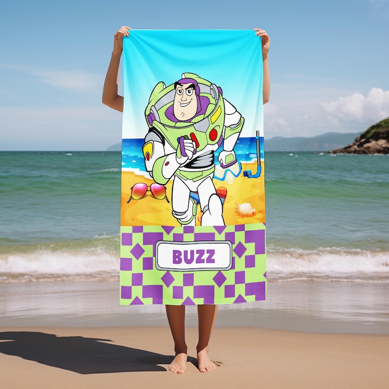 May include: A rectangular beach towel featuring a graphic of the character Buzz Lightyear on a sandy beach with a blue sky. The towel includes a snorkel, sunglasses, and a seashell. The bottom section has a purple and green checkered pattern with a white rectangular label containing the text BUZZ in purple capital letters.