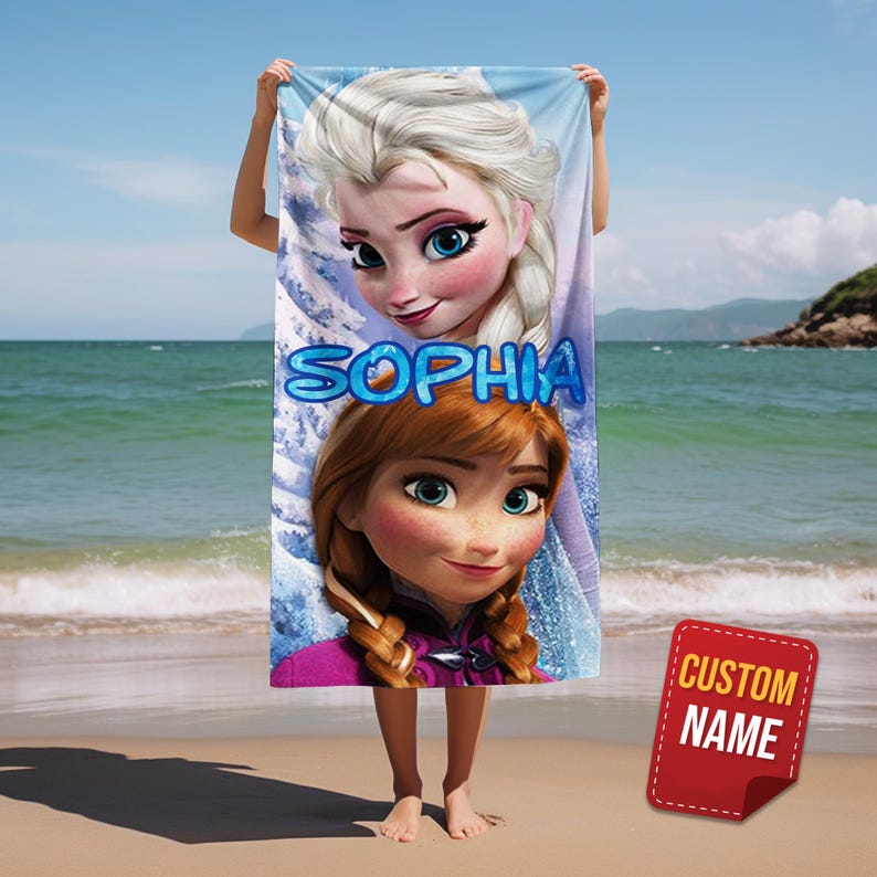 Pode incluir: Uma toalha de praia com Elsa e Anna de Frozen, com o nome "SOPHIA" em letras azuis de bolhas. A toalha &eacute; segurada em uma praia com o oceano ao fundo. Uma etiqueta vermelha com "CUSTOM NAME" est&aacute; no canto inferior direito.