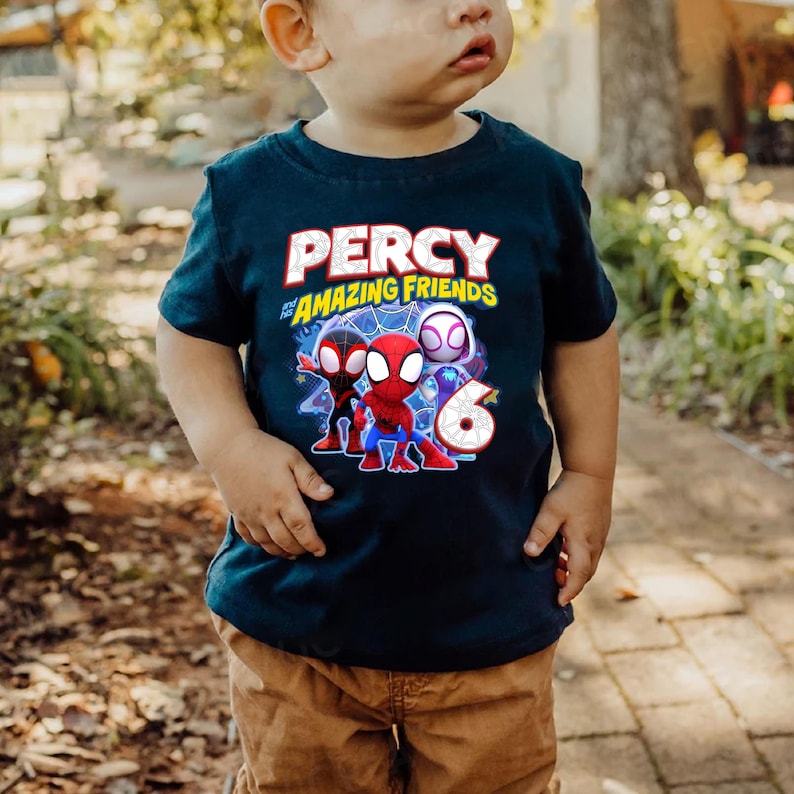 May include: A dark blue short-sleeved t-shirt featuring a graphic print with the text PERCY and AMAZING FRIENDS. The design includes three cartoon spider-themed characters and a large number six with a spiderweb pattern on the front center.