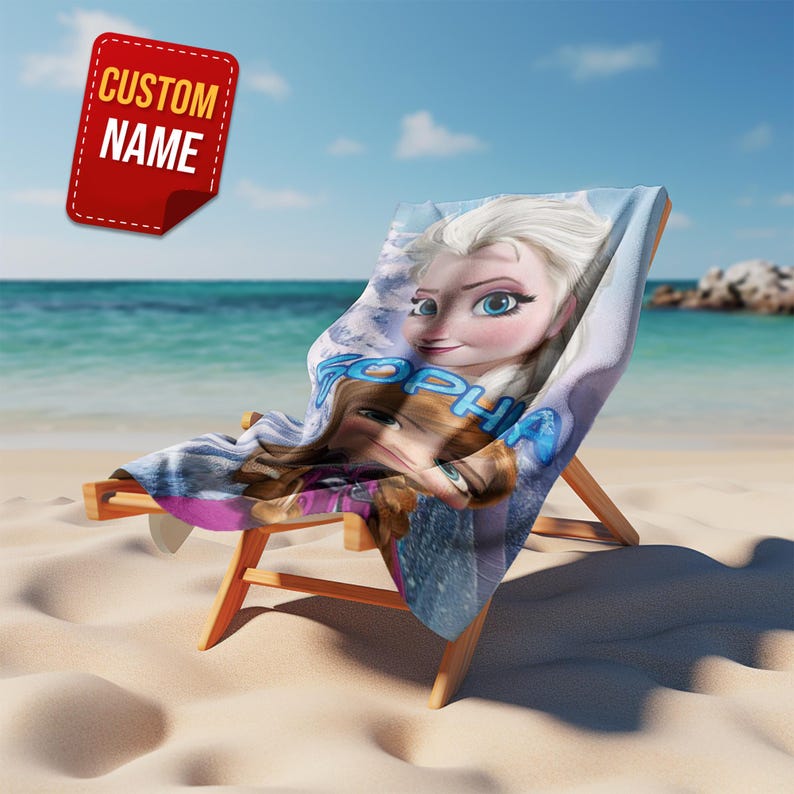 Pode incluir: Uma cadeira de praia com uma etiqueta de nome personalizada e uma toalha de praia com as personagens Elsa e Anna do filme Frozen. A toalha est&aacute; sobre a cadeira de madeira numa praia de areia com o oceano ao fundo.