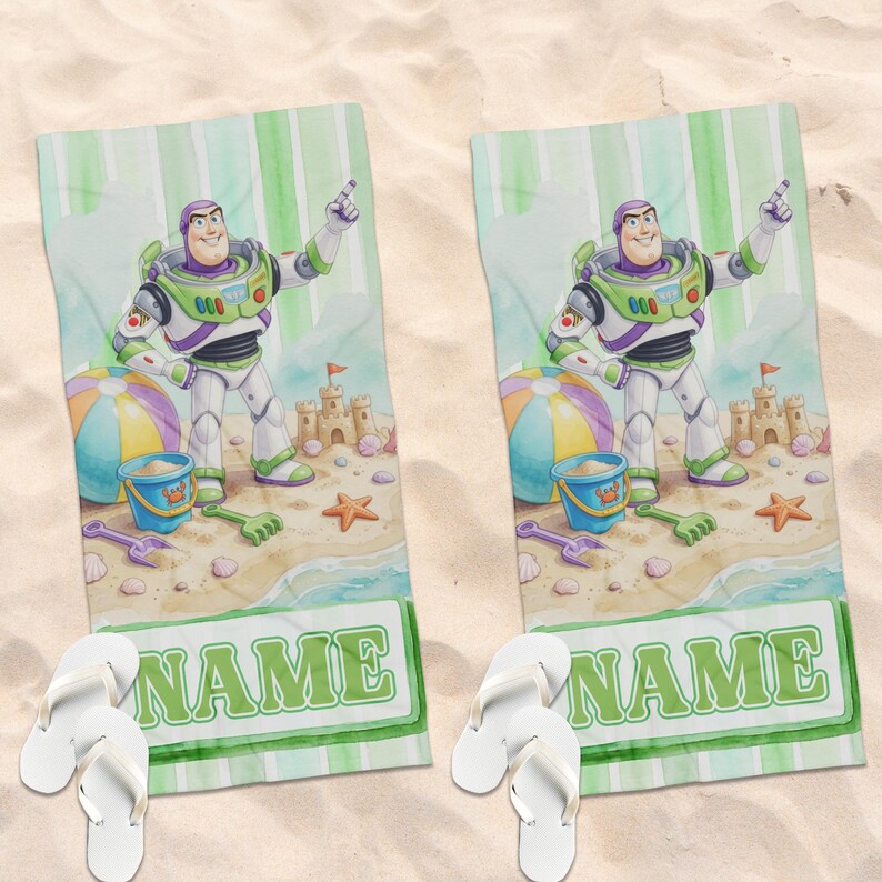 May include: Two rectangular beach towels featuring a cartoon space ranger character in a white, green, and purple suit. The design includes a sandcastle, a beach ball, a blue bucket, and the word NAME in green letters, paired with white flip-flops.