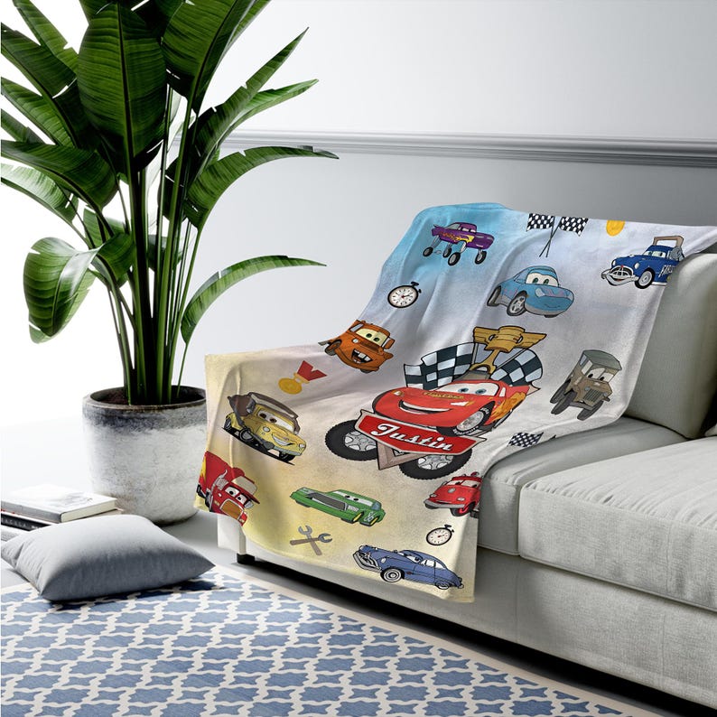 May include: A soft throw blanket featuring a colorful cartoon car print with various vehicles, checkered flags, stopwatches, and the name Justin written in white script on a red bumper. The background is a light gradient of blue and cream.