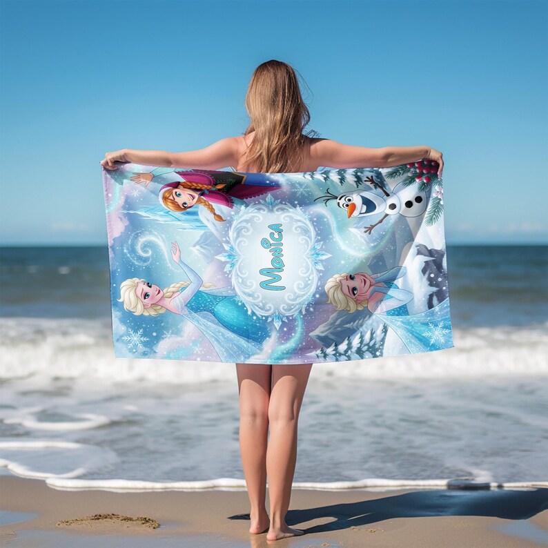 K&ouml;nnte beinhalten: Ein rechteckiges Strandtuch mit Figuren aus dem Film Frozen, darunter Elsa, Anna und Olaf, in einer winterlichen Szene. Das Handtuch hat einen hellblauen Hintergrund mit dem Namen "Monica" in einer dekorativen Schriftart.