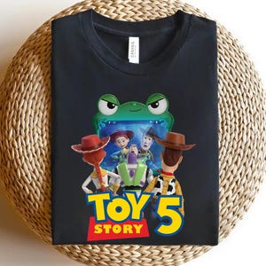 May include: Black t-shirt featuring characters from the animated film Toy Story 5. The design includes Woody, Buzz Lightyear, Jessie, and other characters, with the film's title in yellow and red. The shirt is displayed on a woven surface.