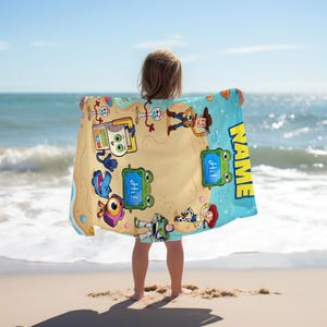 May include: A beach towel featuring cartoon characters on a sandy beach design with a blue border. The towel has the word "NAME" in yellow and the word "Hi!" in blue speech bubbles. The towel is held by a person on a sandy beach with the ocean in the background.