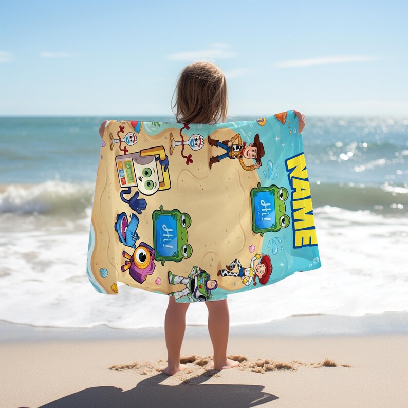 May include: A beach towel featuring cartoon characters on a sandy beach design with a blue border. The towel has the word "NAME" in yellow and the word "Hi!" in blue speech bubbles. The towel is held by a person on a sandy beach with the ocean in the background.