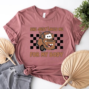 Puede incluir: Una camiseta de color malva con un gr&aacute;fico de una gr&uacute;a marr&oacute;n con auriculares, situada frente a un patr&oacute;n de cuadros blancos y negros. El texto "SHE JUST LIKES ME FOR MY BODY" est&aacute; impreso en letras marrones sobre y bajo el gr&aacute;fico.