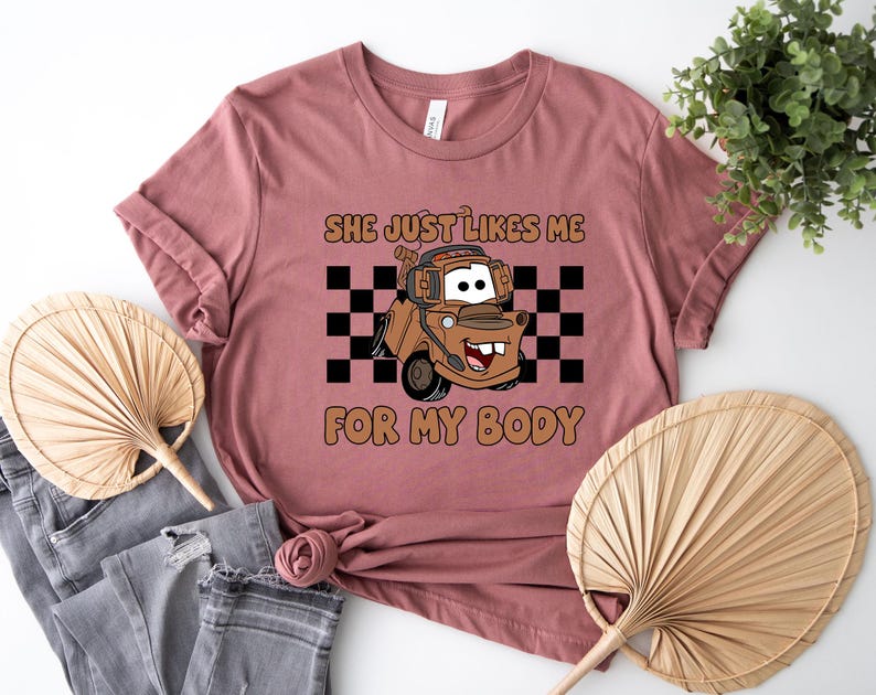 Puede incluir: Una camiseta de color malva con un gr&aacute;fico de una gr&uacute;a marr&oacute;n con auriculares, situada frente a un patr&oacute;n de cuadros blancos y negros. El texto "SHE JUST LIKES ME FOR MY BODY" est&aacute; impreso en letras marrones sobre y bajo el gr&aacute;fico.