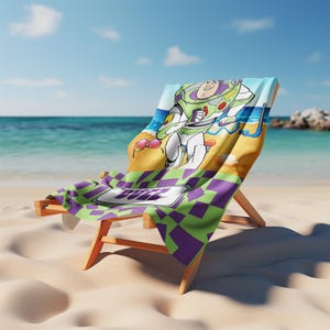 May include: A beach towel draped over a wooden lounge chair featuring a graphic of Buzz Lightyear on a sandy beach with a blue ocean background. The towel has a purple and green checkered pattern at the bottom with the name BUZZ printed in white.