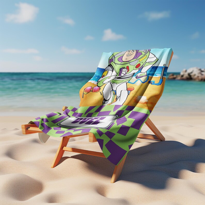 May include: A beach towel draped over a wooden lounge chair featuring a graphic of Buzz Lightyear on a sandy beach with a blue ocean background. The towel has a purple and green checkered pattern at the bottom with the name BUZZ printed in white.