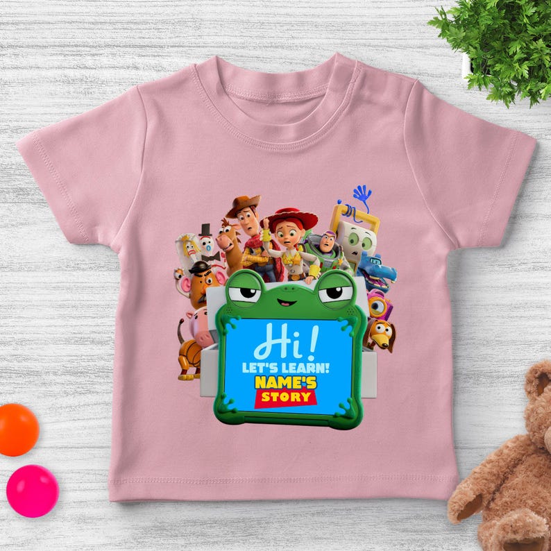 May include: Pale pink short-sleeved t-shirt with a vibrant graphic of animated characters. A green frog-shaped frame displays the word "Hi!" and the phrases "LET'S LEARN! NAME'S STORY". The shirt is designed for children.