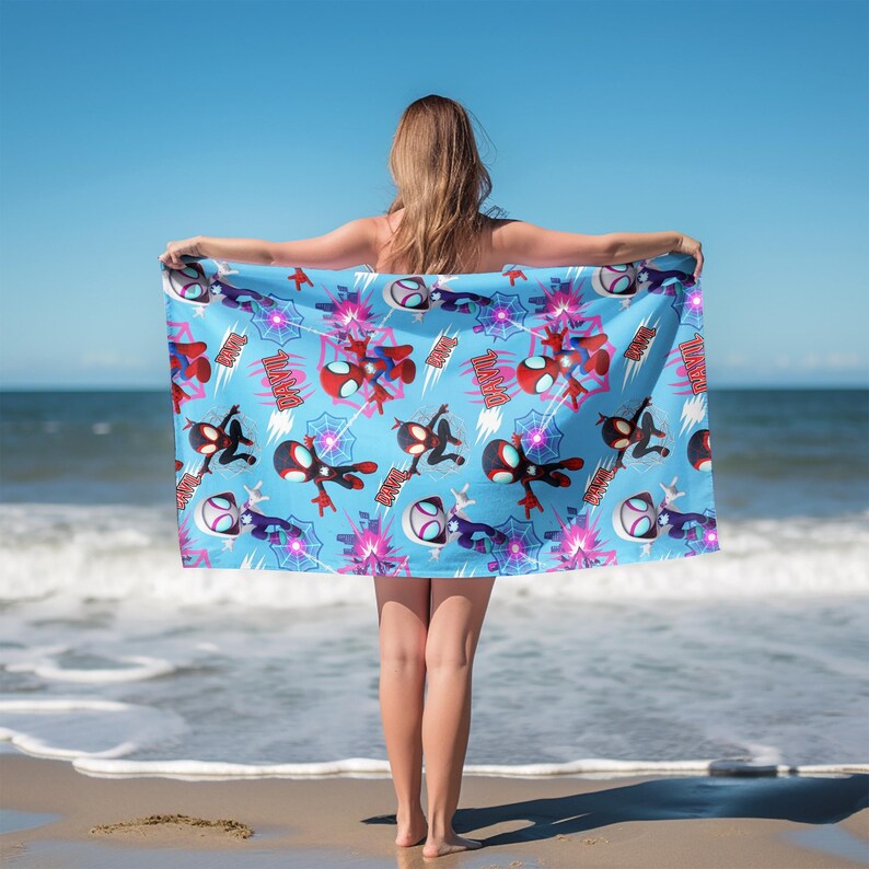 Puede incluir: Una toalla de playa rectangular con fondo azul claro y un patr&oacute;n repetitivo de superh&eacute;roes de dibujos animados en trajes rojos, negros y blancos, telara&ntilde;as y estrellas rosas. La palabra BAM est&aacute; impresa en texto rojo por todo el dise&ntilde;o.
