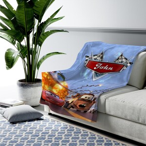 May include: A soft blue throw blanket featuring a graphic print of the characters Lightning McQueen and Mater from the movie Cars. The design includes two checkered racing flags and a red emblem with the name John printed in white script.