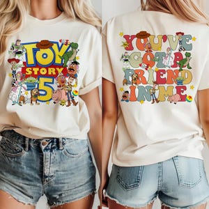 K&ouml;nnte beinhalten: Cremefarbenes T-Shirt mit dem Toy Story 5 Logo auf der Vorderseite und dem Schriftzug "You've Got a Friend in Me" auf der R&uuml;ckseite. Das Design zeigt verschiedene Toy Story Charaktere in einem bunten Cartoon-Stil.