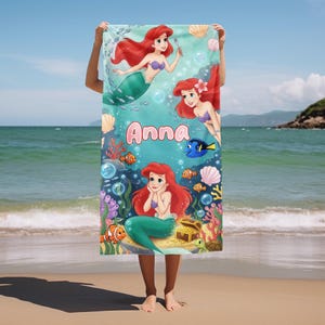 Peut inclure: Une serviette de plage rectangulaire avec une illustration de sir&egrave;ne aux cheveux roux et une queue verte, entour&eacute;e de cr&eacute;atures marines. Le nom "Anna" est &eacute;crit en rose sur la serviette.