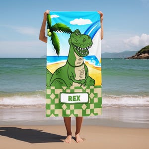 May include: A rectangular beach towel featuring a cartoon green Tyrannosaurus Rex standing on a sandy beach with a palm tree and blue sky. The lower section has a green and beige geometric pattern with a white rectangular label containing the text REX.