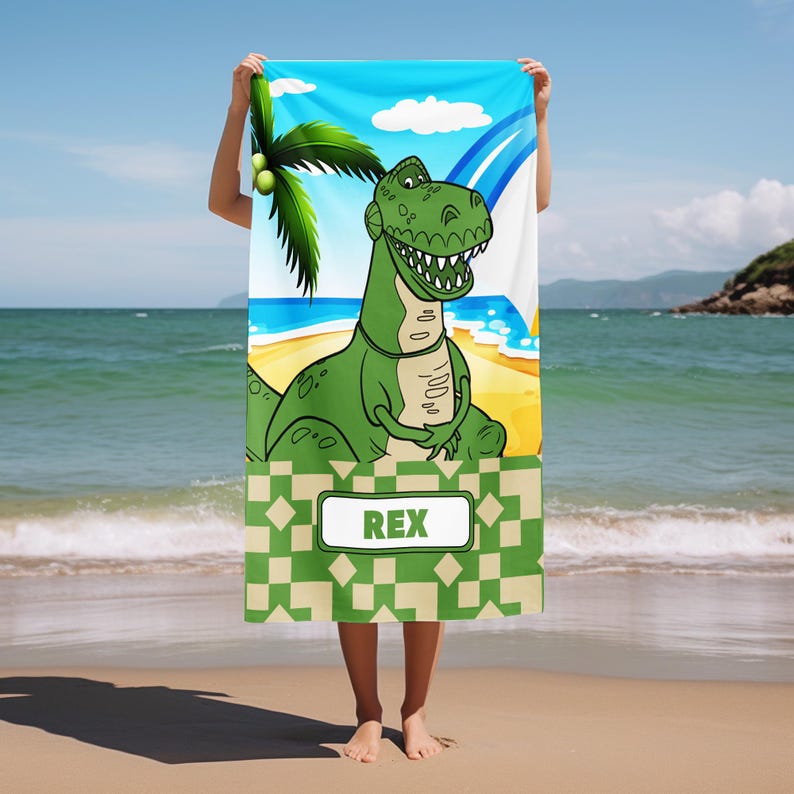 May include: A rectangular beach towel featuring a cartoon green Tyrannosaurus Rex standing on a sandy beach with a palm tree and blue sky. The lower section has a green and beige geometric pattern with a white rectangular label containing the text REX.