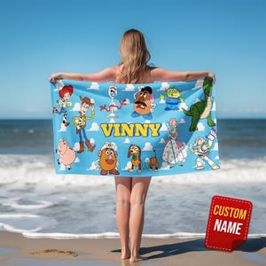 May include: A blue beach towel featuring various Toy Story characters and the name "VINNY" in yellow block letters. The towel is held up by a person on a beach with the ocean and sky in the background. A red tag with "CUSTOM NAME" is in the bottom right corner.