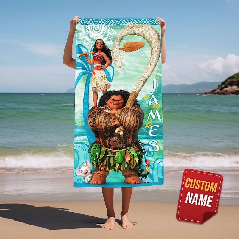 May include: A beach towel featuring characters from the animated movie Moana, including Maui, Moana, and Pua. The towel has a blue and turquoise background with the name "JAMES" printed vertically. A red tag with "CUSTOM NAME" is in the lower right corner.