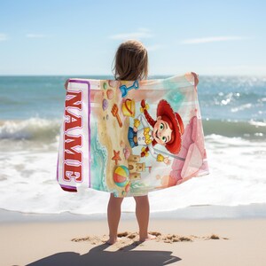 Puede incluir: Una toalla de playa rectangular con un personaje de vaquera de dibujos animados con trenzas rojas, un castillo de arena, una pelota de playa y una pala sobre la arena. La toalla incluye una franja blanca vertical con el texto NAME en rojo.