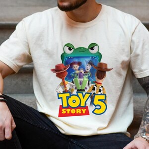May include: Cream-colored t-shirt featuring a colorful graphic from the movie Toy Story 5. The design includes characters like Woody, Buzz Lightyear, and Jessie, with the text "TOY STORY 5" in bold, colorful letters. The shirt is made of a soft material.