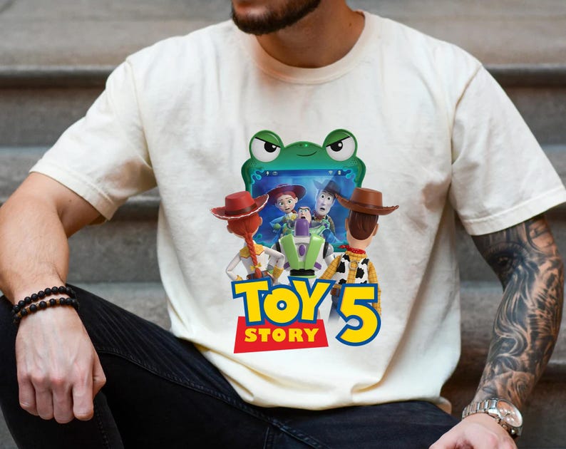 May include: Cream-colored t-shirt featuring a colorful graphic from the movie Toy Story 5. The design includes characters like Woody, Buzz Lightyear, and Jessie, with the text "TOY STORY 5" in bold, colorful letters. The shirt is made of a soft material.