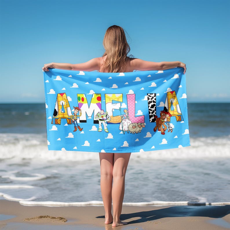May include: A rectangular beach towel featuring a light blue background with white clouds. The name AMELIA is printed in large, colorful, patterned letters. Various animated characters are positioned in front of the letters on the towel.