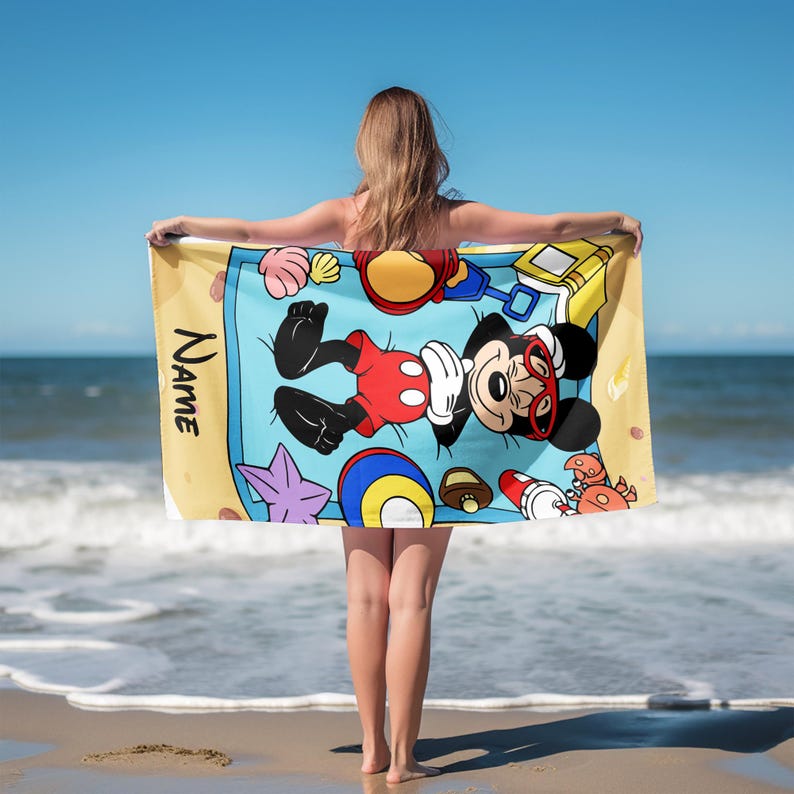 May include: Beach towel featuring Mickey Mouse in sunglasses, lying on a blue beach blanket with sand toys and seashells. The towel has a yellow border and the word "Name" printed vertically on the side.