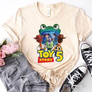 May include: Cream-colored t-shirt featuring a colorful graphic from the movie Toy Story 5. The graphic includes characters like Woody, Buzz Lightyear, and Jessie. The shirt is paired with gray jeans and black sneakers.