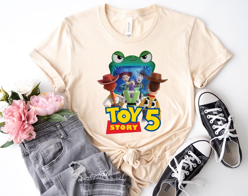 May include: Cream-colored t-shirt featuring a colorful graphic from the movie Toy Story 5. The graphic includes characters like Woody, Buzz Lightyear, and Jessie. The shirt is paired with gray jeans and black sneakers.