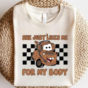 Puede incluir: Una camiseta de color crema con un gr&aacute;fico de una gr&uacute;a marr&oacute;n que lleva auriculares. Detr&aacute;s del cami&oacute;n hay un patr&oacute;n de cuadros blancos y negros. El texto "SHE JUST LIKES ME FOR MY BODY" aparece en letras marrones, redondeadas y en negrita.