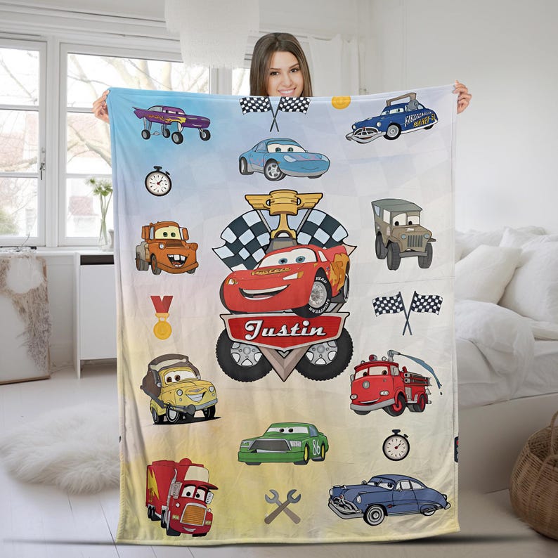 May include: A soft fleece blanket featuring a colorful print of various cartoon race cars, including a red car with the name Justin printed on a banner, checkered flags, a gold trophy, stopwatches, and tools against a light blue and white background.
