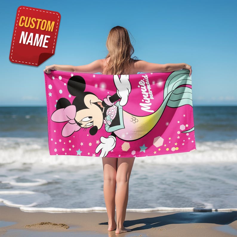 Pode incluir: Uma toalha de praia rosa com Minnie Mouse como sereia, com o texto "Minnie mermaid". A toalha tem uma etiqueta de nome personalizada no canto superior esquerdo. O design inclui estrelas e formas geom&eacute;tricas.