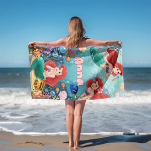 Peut inclure: Une serviette de plage rectangulaire avec une illustration color&eacute;e d'Ariel la Petite Sir&egrave;ne, avec le nom "Anna" en rose. Le motif comprend des cr&eacute;atures marines et des &eacute;l&eacute;ments sous-marins, sur fond bleu.