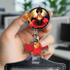 May include: A retractable badge reel featuring a circular acrylic charm of Jafar holding a golden cobra staff, with a secondary dangling charm of Iago the red parrot. The design includes vibrant red, black, yellow, and purple colors on clear acrylic.