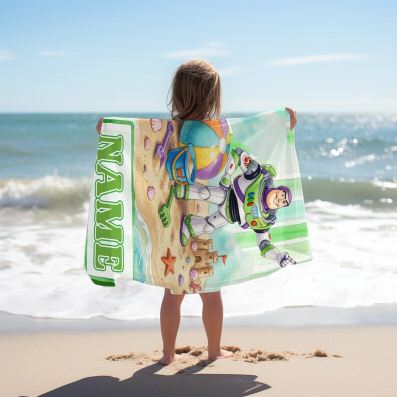 May include: A rectangular beach towel featuring a cartoon illustration of Buzz Lightyear on a sandy beach with a beach ball, a bucket, and a sandcastle. The towel includes a vertical green border with the word NAME printed in white block letters.