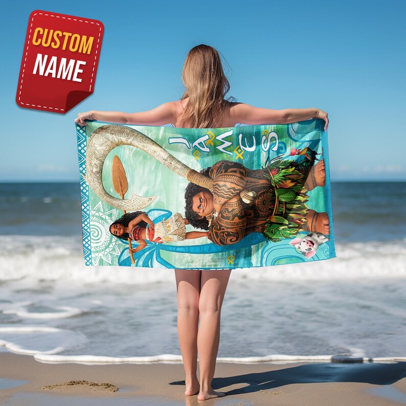 May include: A colourful beach towel with characters from a popular animated film. The towel displays a detailed illustration of a Polynesian demigod, a young woman, and a pig, set against a turquoise and blue background. The towel is held by a person on a sandy beach.