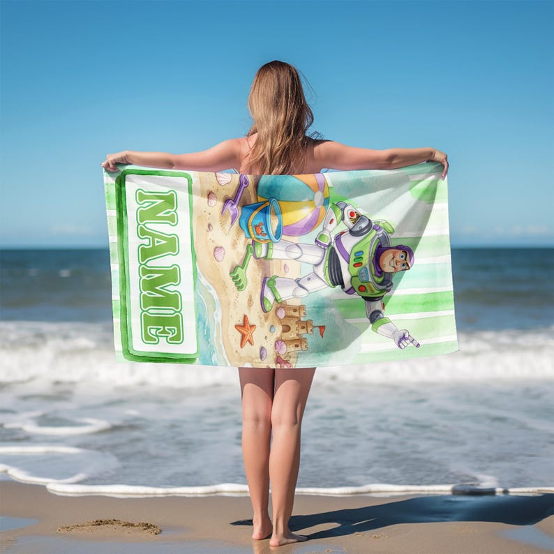 May include: A rectangular beach towel featuring a green and white striped background with a graphic of a space ranger character, a sandcastle, a beach ball, and a bucket. The towel includes a white vertical banner with the word NAME in green.