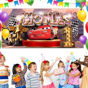 May include: A birthday banner featuring the name "THOMAS" in large, metallic letters, with a red race car and the number "1". The banner is decorated with balloons and checkered flags, suggesting a car-themed first birthday party.