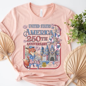 Puede incluir: Una camiseta rosa claro con un gr&aacute;fico que celebra el 250 aniversario de los Estados Unidos de Am&eacute;rica. El dise&ntilde;o incluye un zorro y un conejo de dibujos animados, fuegos artificiales y un castillo. El texto dice "United States of America 250th Anniversary."