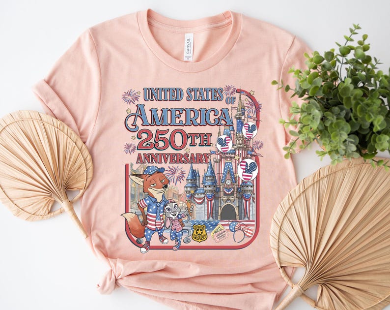 Puede incluir: Una camiseta rosa claro con un gr&aacute;fico que celebra el 250 aniversario de los Estados Unidos de Am&eacute;rica. El dise&ntilde;o incluye un zorro y un conejo de dibujos animados, fuegos artificiales y un castillo. El texto dice "United States of America 250th Anniversary."