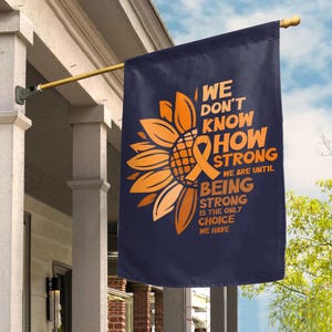 May include: A dark blue rectangular garden flag featuring a large orange sunflower graphic with an integrated awareness ribbon. The text reads: WE DON'T KNOW HOW STRONG WE ARE UNTIL BEING STRONG IS THE ONLY CHOICE WE HAVE, printed in orange.