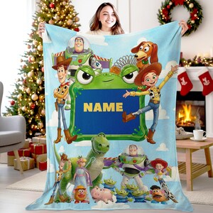 May include: A light blue fleece blanket featuring Toy Story characters including Woody, Buzz Lightyear, Jessie, Rex, Slinky Dog, and the Aliens. A central green frog-shaped frame contains the yellow text NAME on a dark blue background.