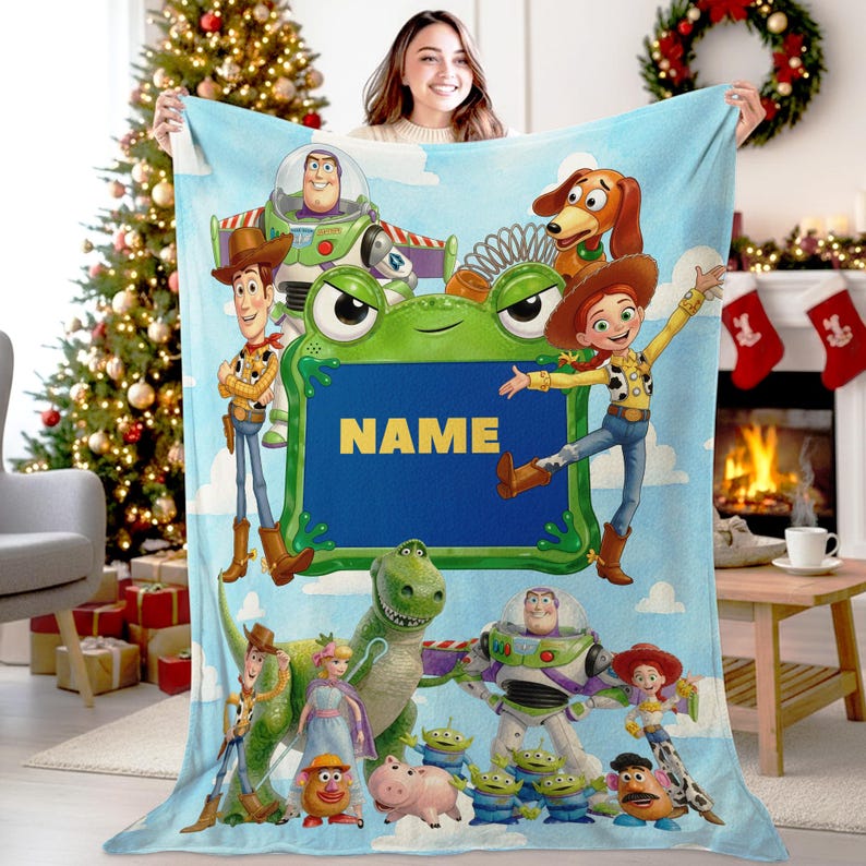 May include: A light blue fleece blanket featuring Toy Story characters including Woody, Buzz Lightyear, Jessie, Rex, Slinky Dog, and the Aliens. A central green frog-shaped frame contains the yellow text NAME on a dark blue background.