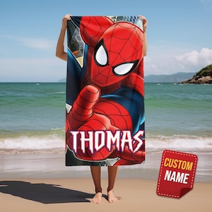 May include: A red and blue Spider-Man beach towel with the name "THOMAS" in white script. The towel features a large graphic of Spider-Man with a "CUSTOM NAME" tag in the corner. The towel is held up on a beach with the ocean in the background.