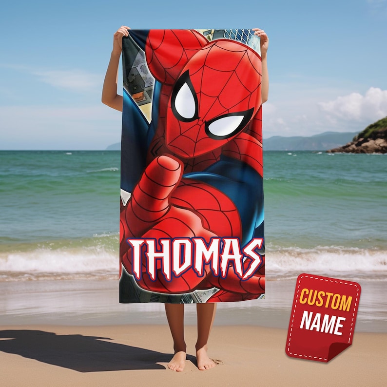 May include: A red and blue Spider-Man beach towel with the name "THOMAS" in white script. The towel features a large graphic of Spider-Man with a "CUSTOM NAME" tag in the corner. The towel is held up on a beach with the ocean in the background.